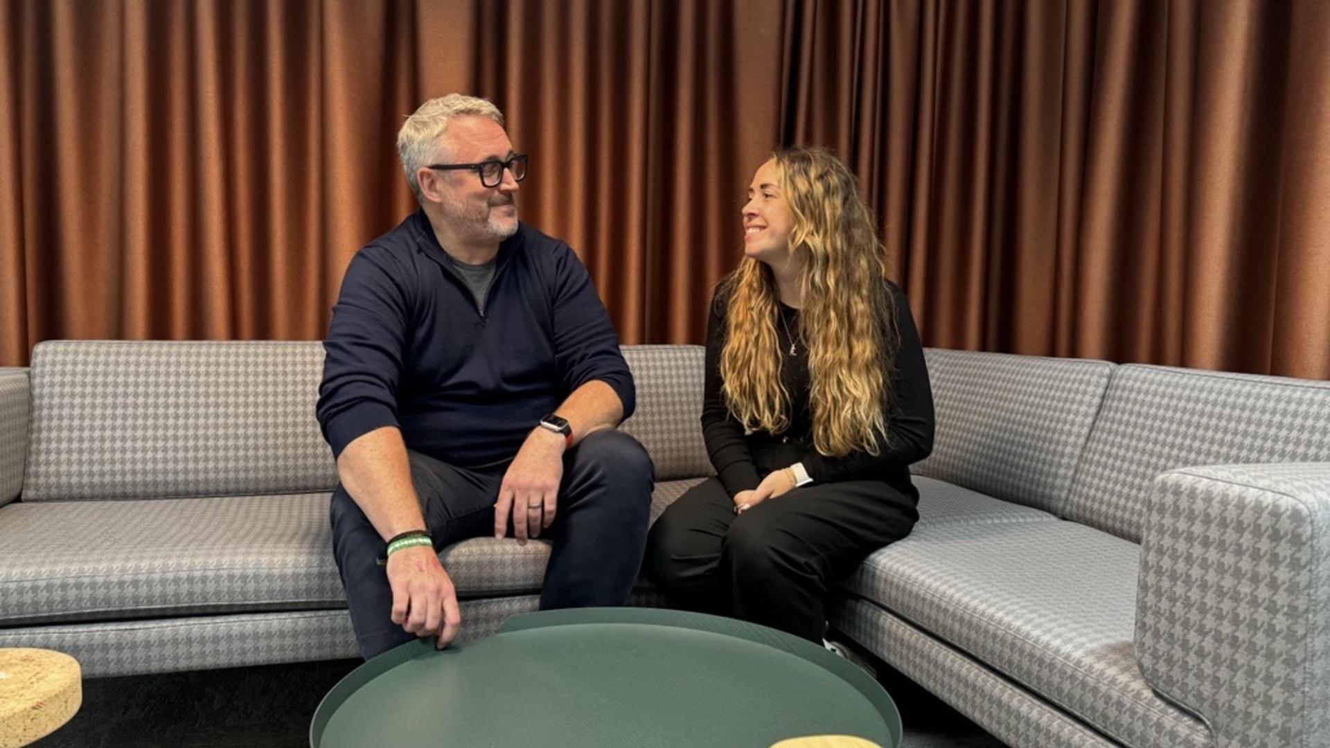 A man named Mike, sitting on a grey, L-shaped couch, with a woman called Lil. They are smiling.