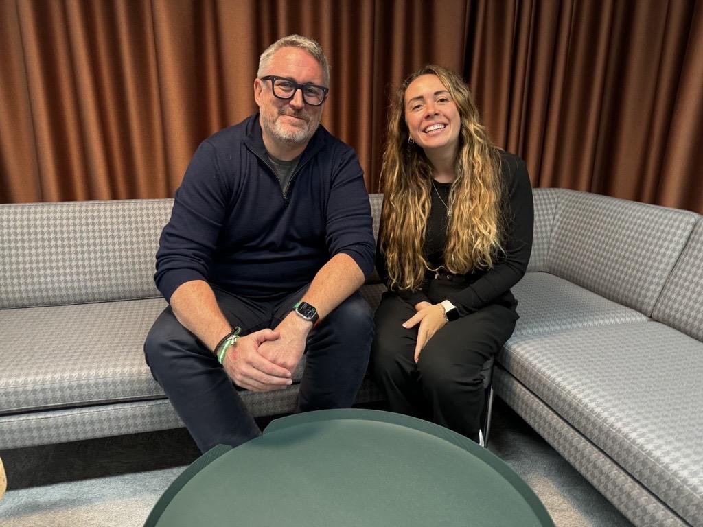 A man named Mike, sitting on a grey, L-shaped couch, with a woman called Lil. They are smiling.